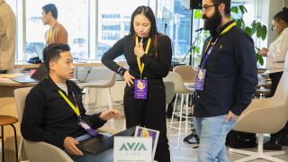 Three people are engaged in a discussion in a bright, modern workspace. One person sits with a laptop while the other two stand nearby, all wearing event badges and lanyards. A table with signage and materials is in the foreground, and other people work in the background.
