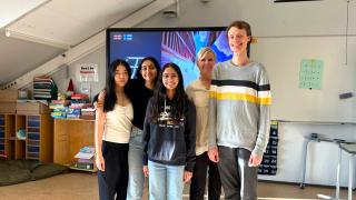 Viisi henkilöä seisoo rivissä luokkahuoneessa. Takana on hyllyjä, kirjoja ja iso näyttö.